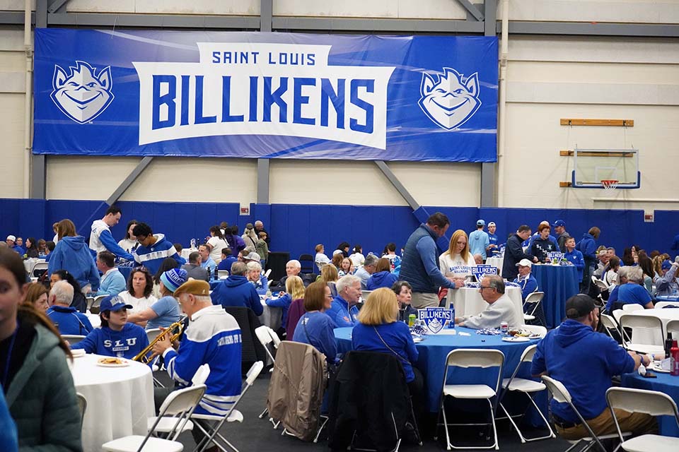 People sit at tables eating and talking. A large banner in the background says Saint Louis Billikens.