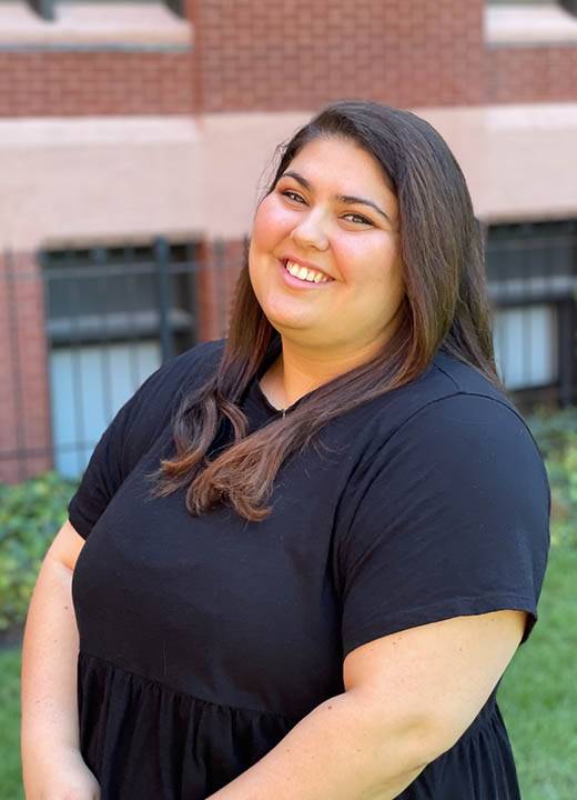 Gabby Sabat, wearing all black, smiles at the camera while standing in front of DuBourg Hall.