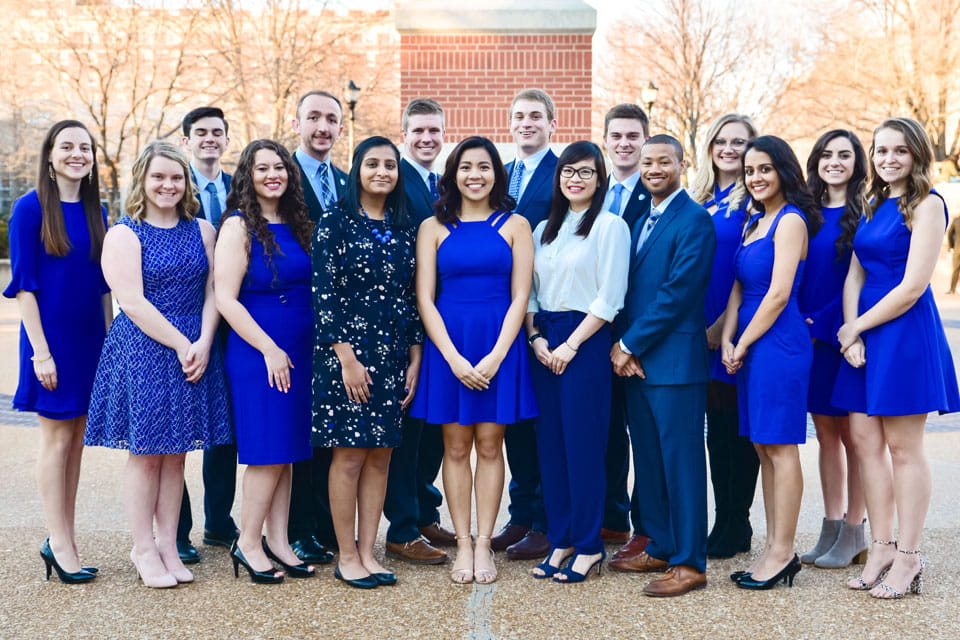 A group of students poses in front of the clock tower, wearing dresses and suits.