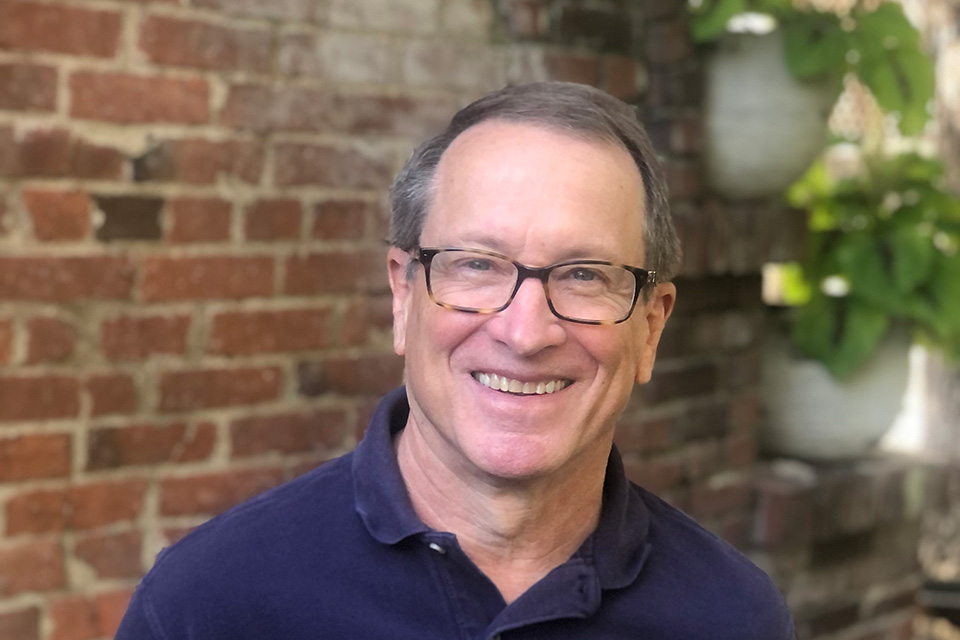 A man poses for a photo with a brick wall and plants behind him.