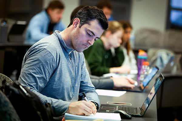 A male student writes in a textbook while sitting in a classroom. His laptop is open in front of him.