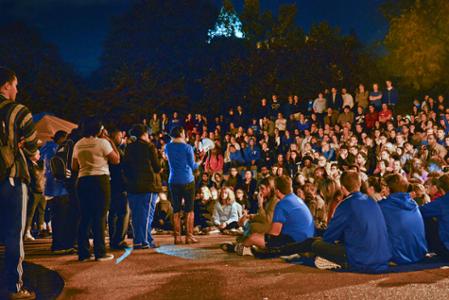 Students crowd around the clock tower at night while a speaker stands at a microphone.