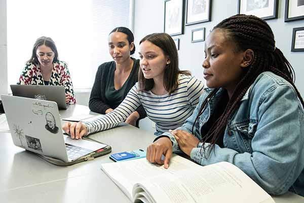 Four SLU students sit at a table in a classroom. Everyone has a laptop or a textbook open in front of them.