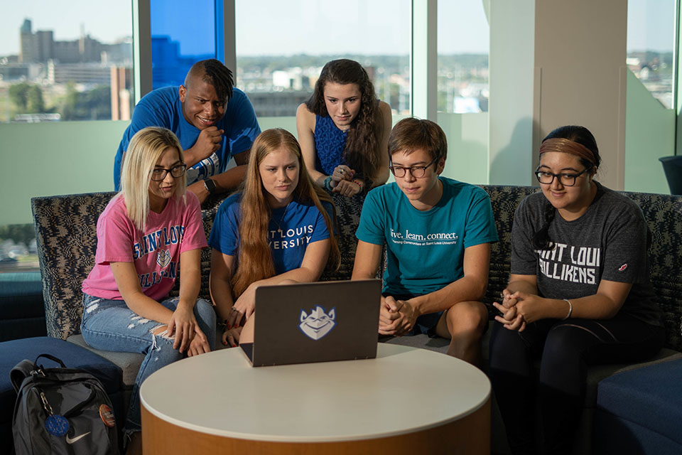 Four students sit on a semi-circular sofa in a residence hall lounge, two stand behind them. All looking at a laptop screen.