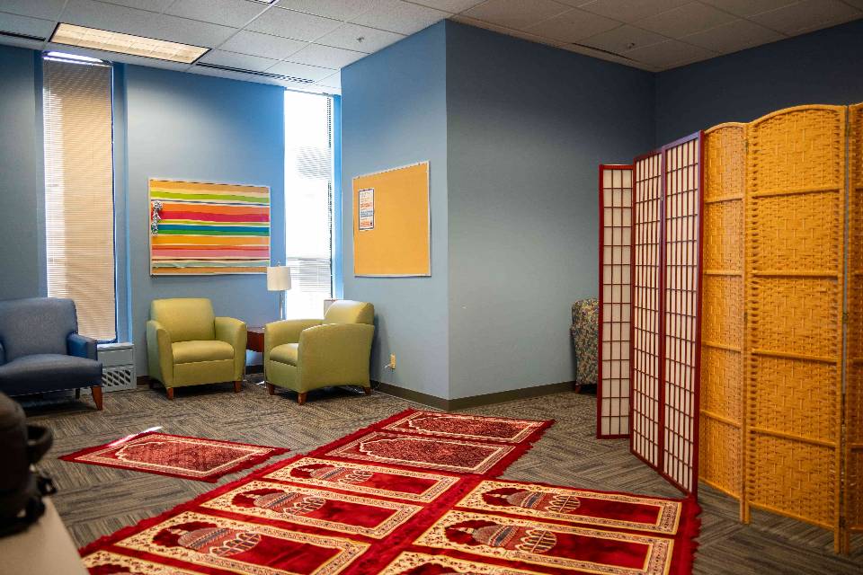 A room with blue walls and comfortable chairs. Prayer rugs are seen lined up on the floor.