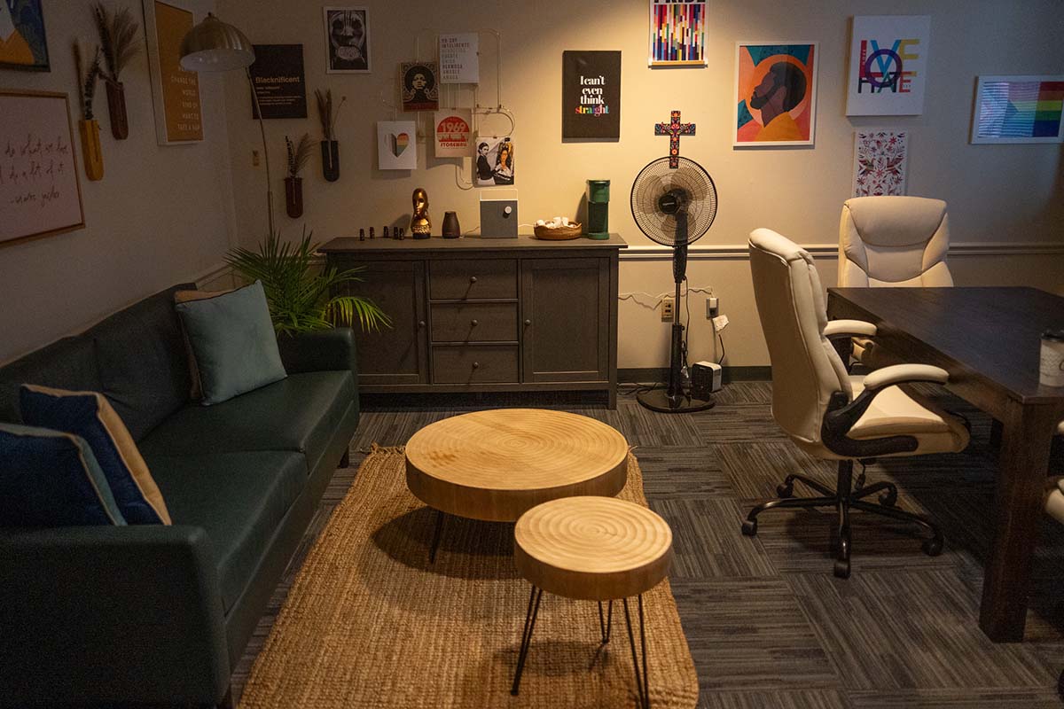 Image of a room with low lighting, a table and chairs, sofa, woven floor rug and round coffee tables, with posters on the wall.