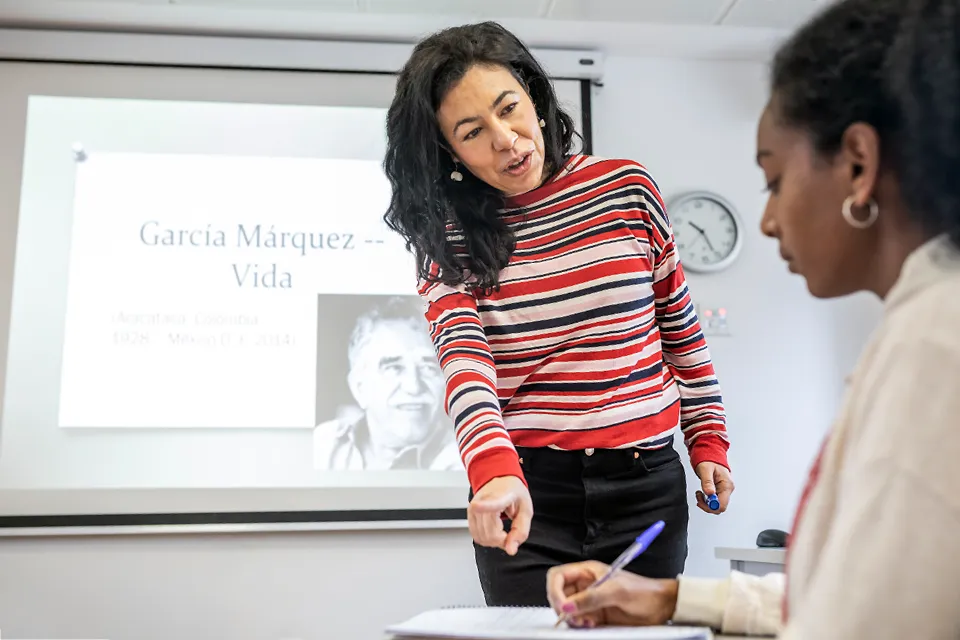 A standing teacher points to the notebook of a student seated at her desk in the classroom. The screen behind has information about Gabriel Garcia Marquez.