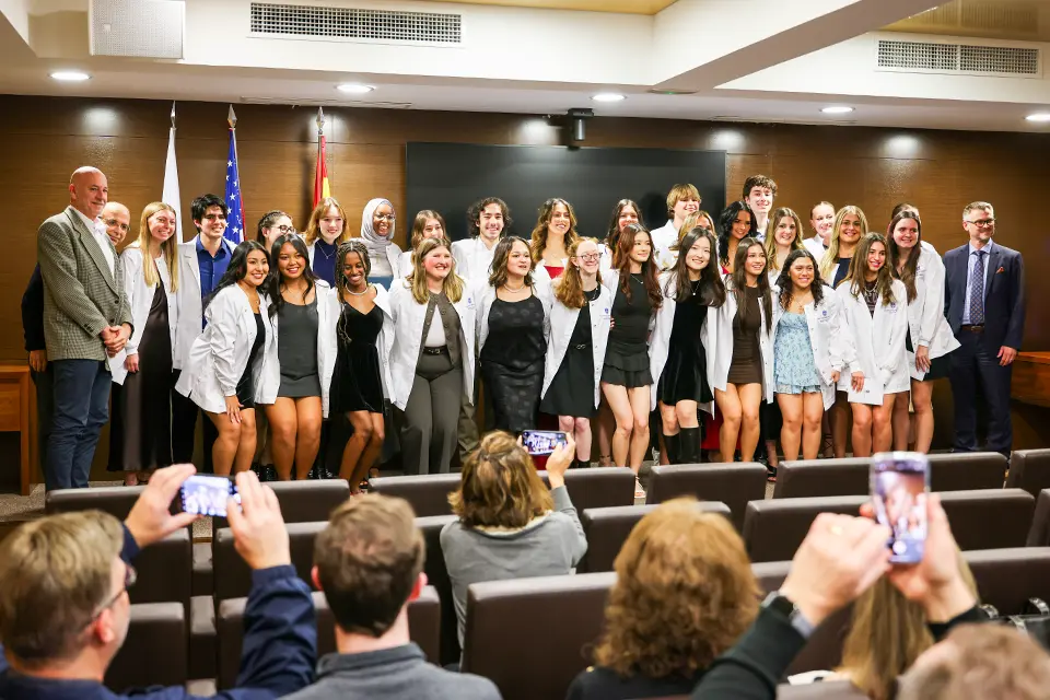 A group of students wearing white coats with professors on the stage, while the audience takes some shots of them.