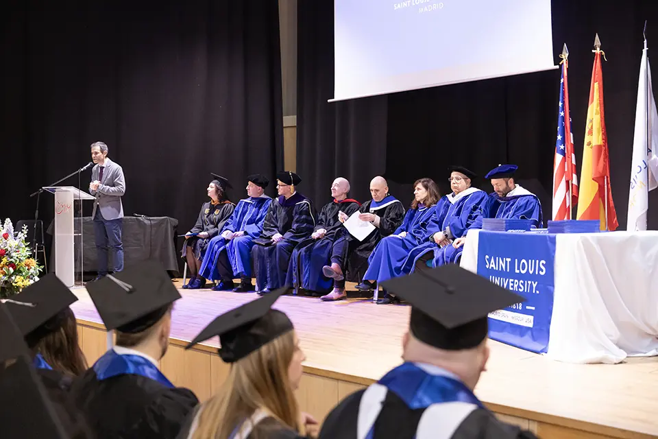 Man speaks on stage in front of podium to an auditorium full of graduating students and other attendees present.