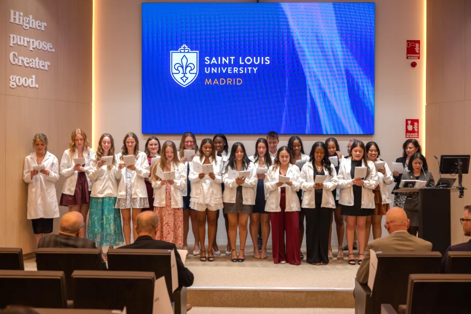 Students with white coats on are reading off pamphlets together in front of an audience. The logo of the University is on the screen behind them. A sign on the wall reads "Higher purpose. Greater good."