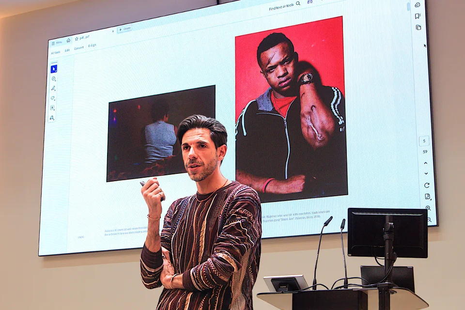 Photojournalist Francesco Bellina speaks in an auditorium, gesturing while presenting images from his "Oriri" project on a large screen behind him, including portraits related to migration and human trafficking.