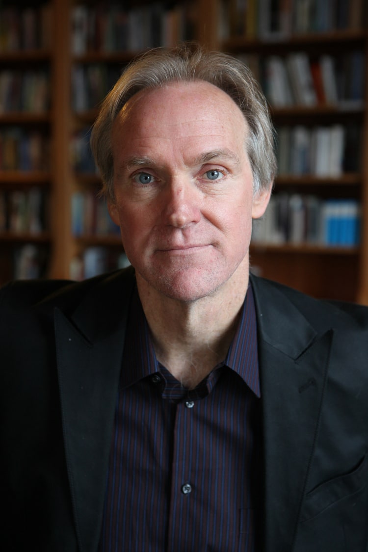 Poet Tom Sleigh sits in a library room with books on shelves behind him