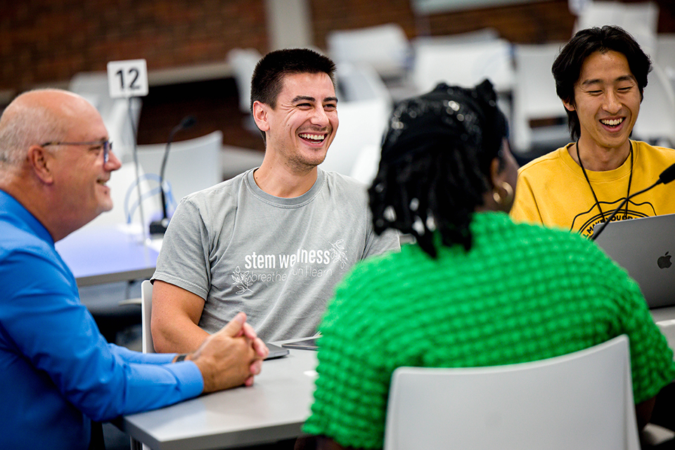 A professor sits at one end of a table while students discuss and collaborate.