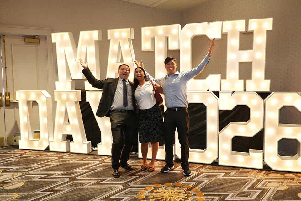 Three students pose for a photo in front of a large lit display that reads "Match Day 2026"