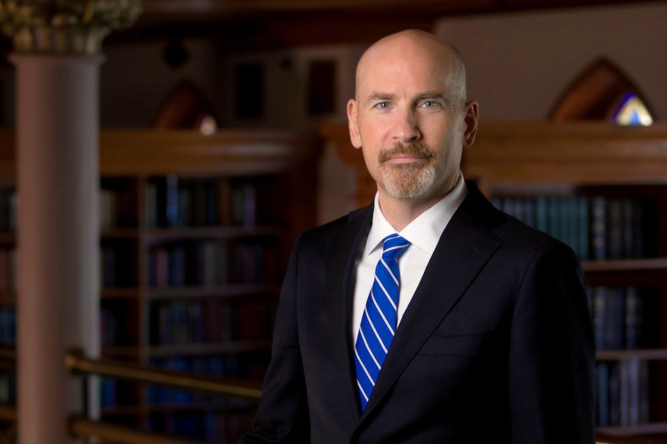 SLU President-Elect Edward J. Feser, wearing a dark suit and royal blue tie, stands before a collection of bookshelves, showcasing a variety of books.