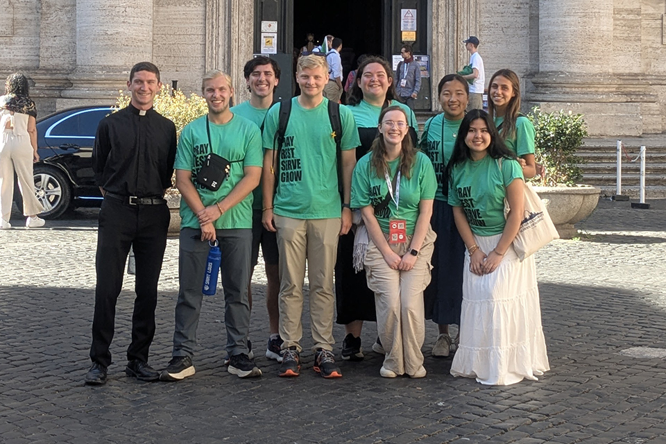 Students wearing green shirts and a priest smile for a photo outdoors.