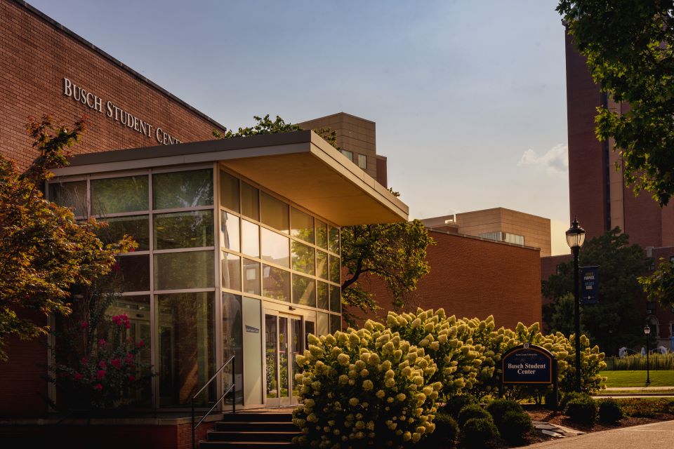 Outside of a building surrounded by bushes and trees on SLU's campus at sunset. A blue sign reads  "Busch Student Center".