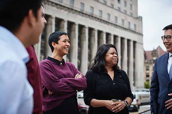 A group of five people converse while standing outside in front of a large building.