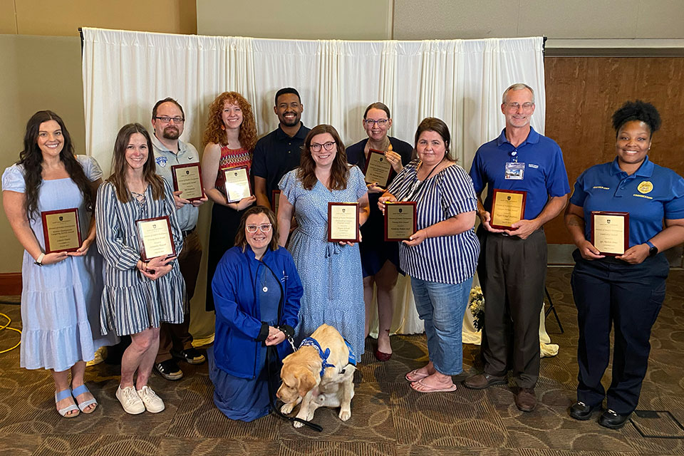 A group of award winners pose for a photo, each holding a plaque. Woody, the dog, is also in the picture, but is not holding a plaque.