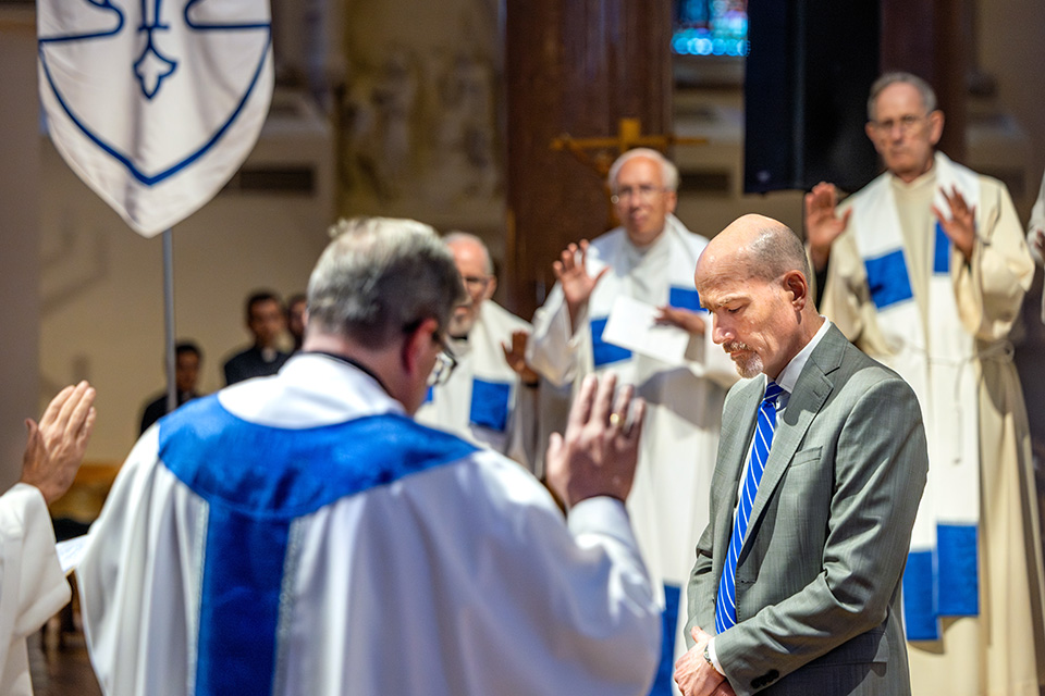 Edward Feser at his missioning as SLU's president A bald white man in a grey suit stands in the middle of a group of Catholic priests in white garb.