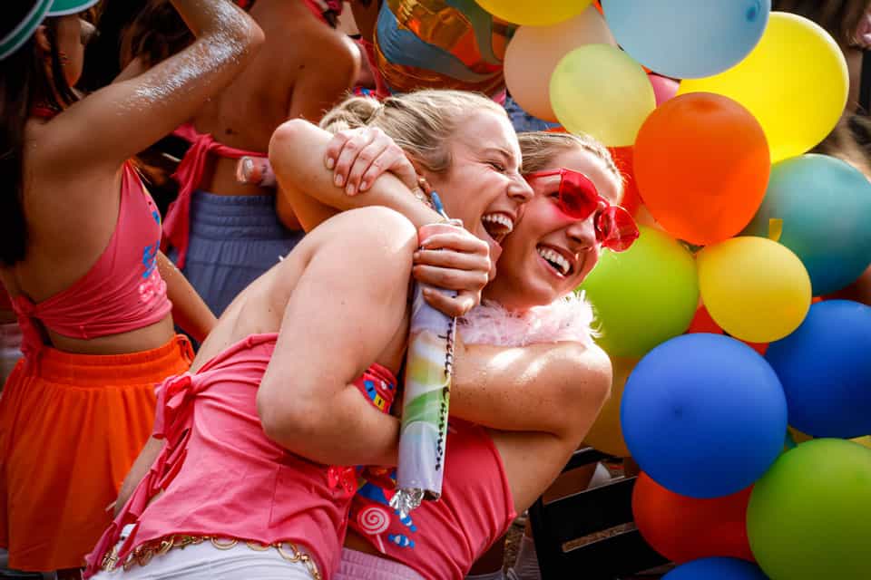 Two girls embrace with a hug in front of assorted colored balloons